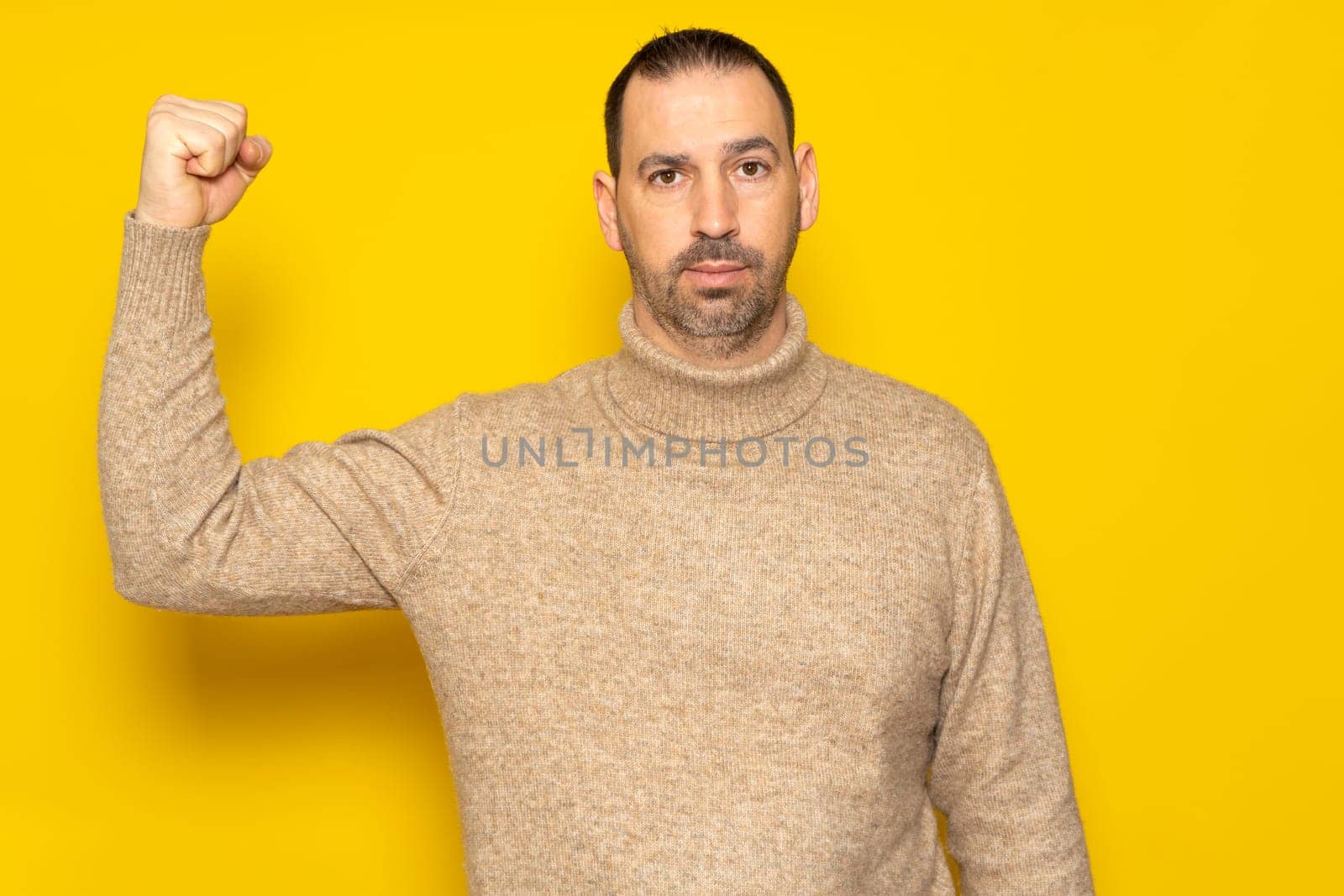 Bearded Hispanic man in his 40s wearing a beige turtleneck showing off his biceps exposing his sorry state of shape, isolated on yellow studio background