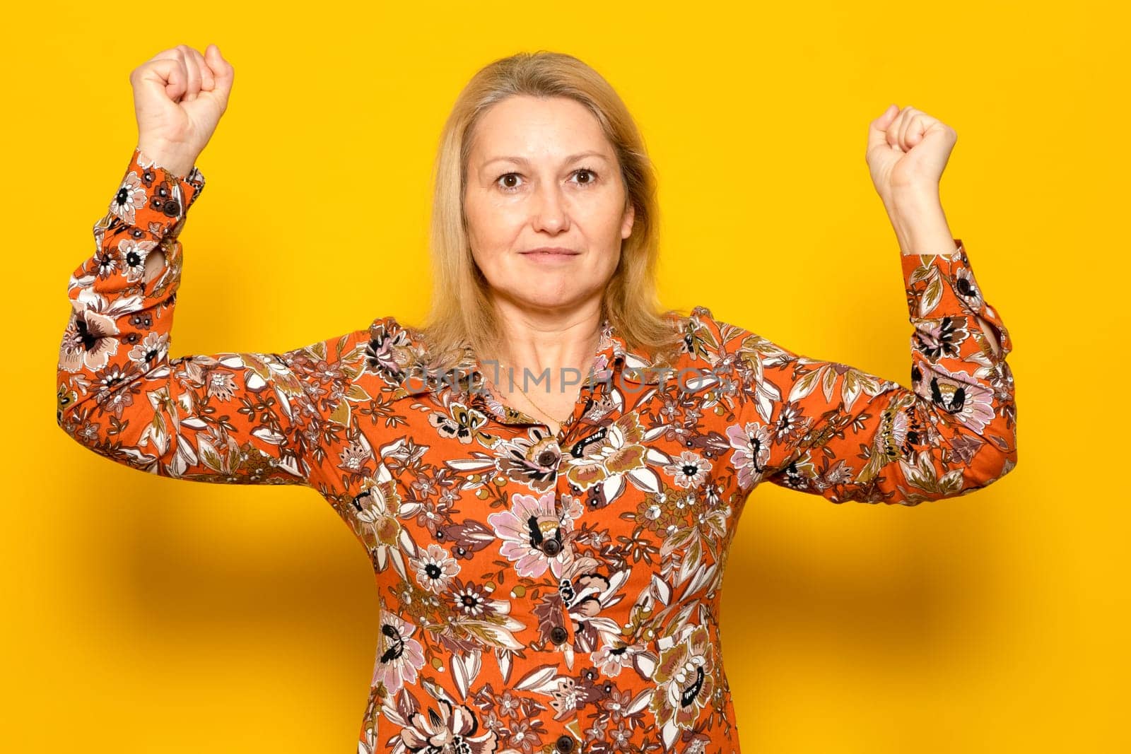 Strong and powerful caucasian woman, raises her arms and shows biceps, wears a patterned shirt, pattern on a yellow background. look at my muscles