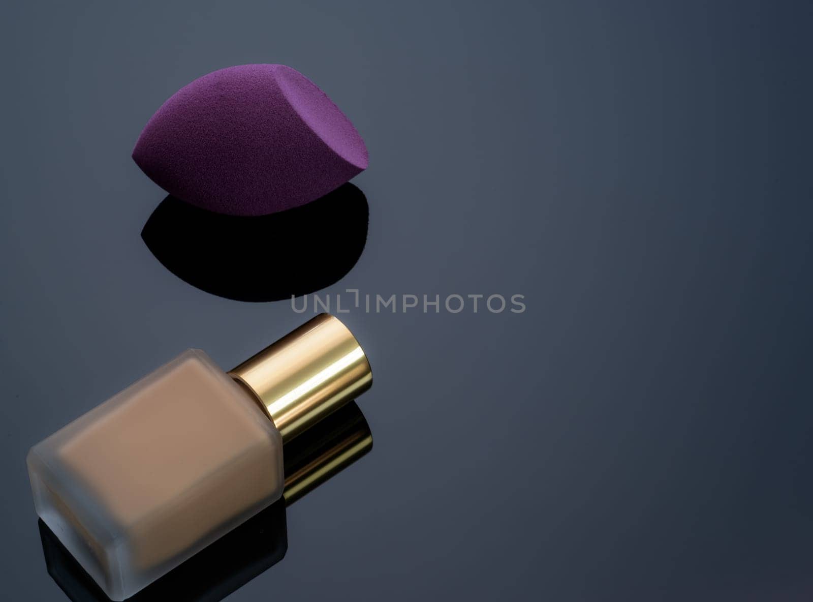 Purple makeup sponge on dark background. Soft makeup cosmetic sponge. Flat-ended makeup sponge with blur makeup foundation bottle. Liquid beige make-up foundation and beauty blender. Cosmetic product. by Fahroni