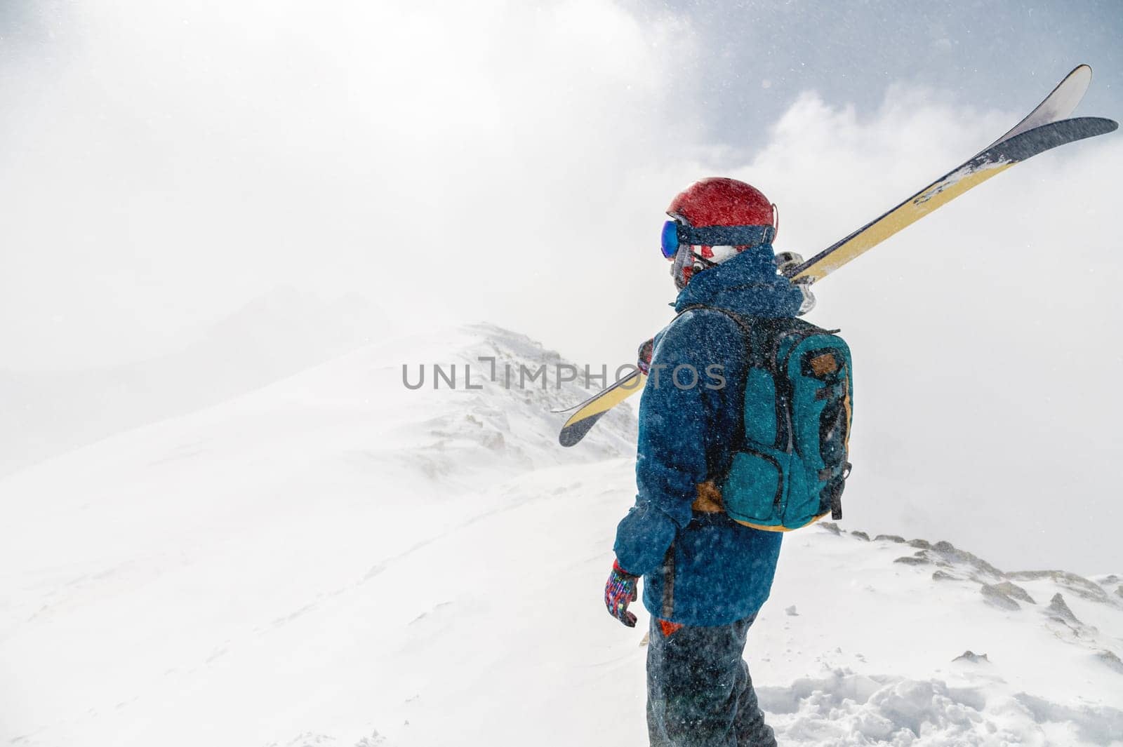 a man stands with his back to the camera with alpine skis on his shoulder against the backdrop of a mountain during a snowfall.