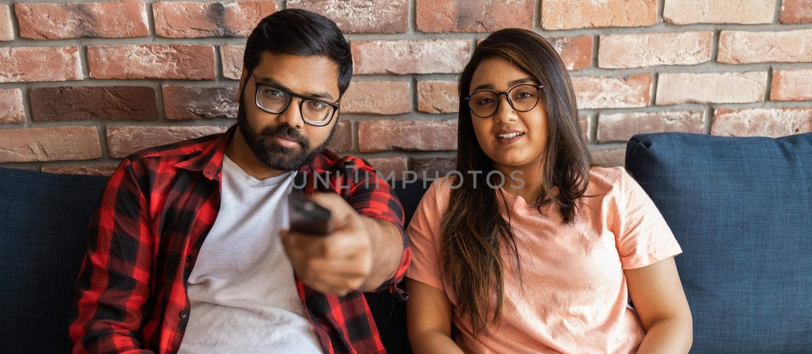 Happy indian or latino couple sitting on sofa and watching action movie on TV or criminal blockbuster on streaming service and talking and discussing acting. Girlfriend and boyfriend resting at home in evening