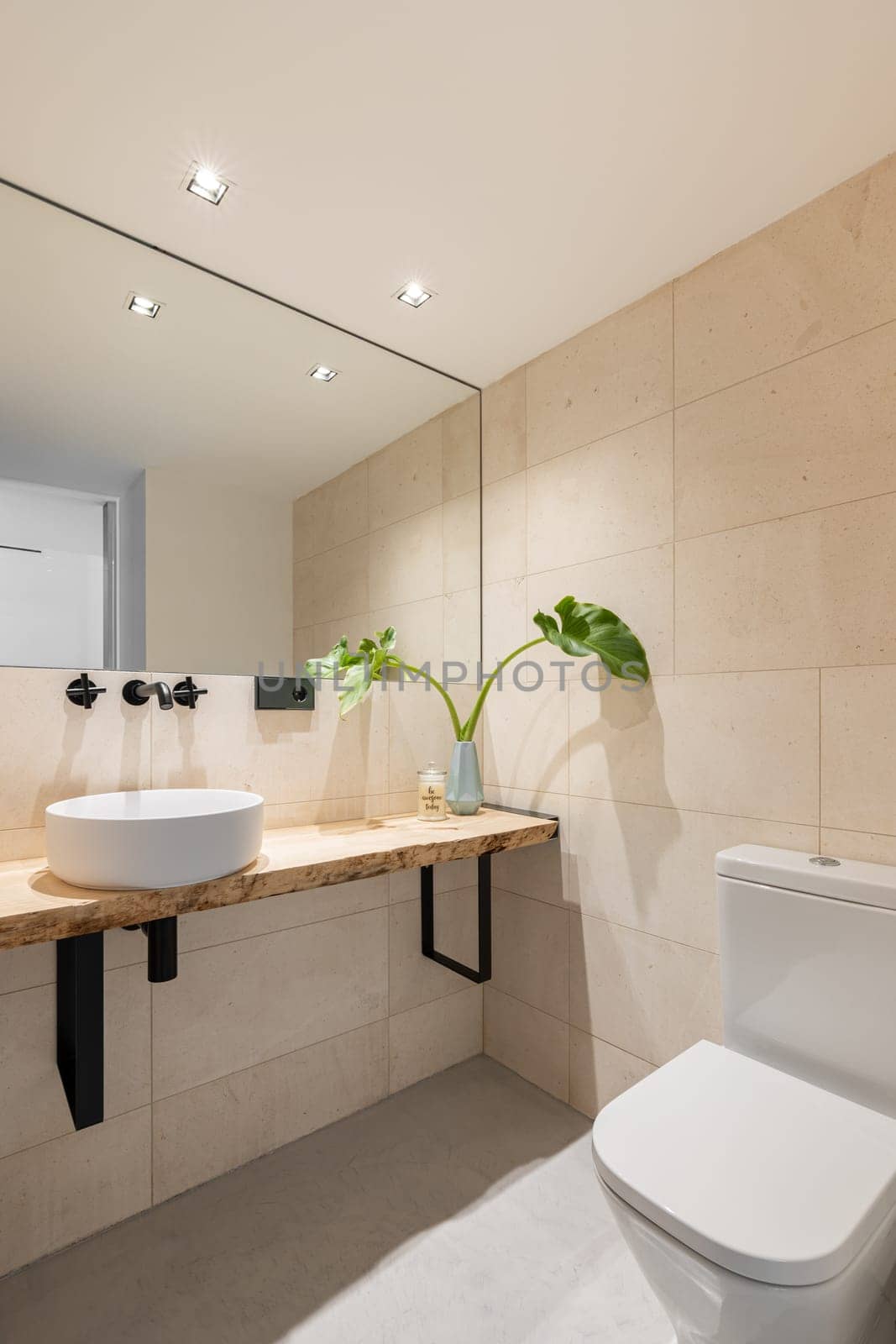 Vertical shot of a bright bathroom with beige tiles on the walls and a large mirror with a sink in a wooden countertop and a toilet bowl. The concept of renovation in an old apartment by apavlin