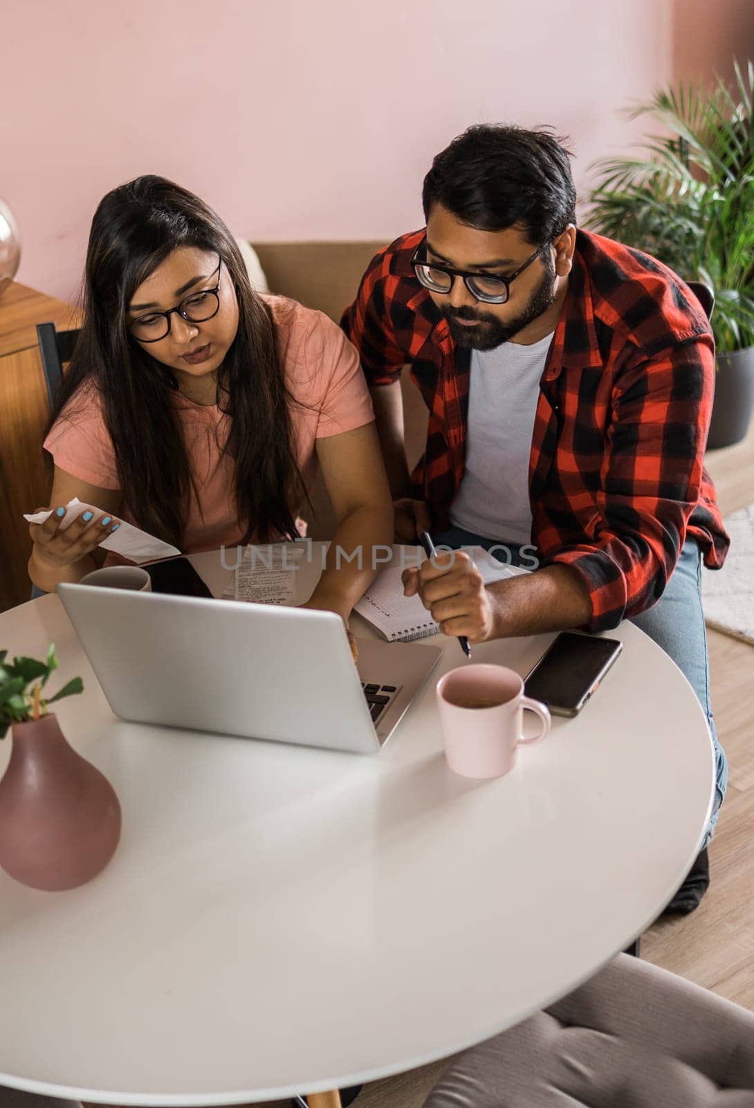 Serious wife and husband planning budget, checking finances, focused young woman using online calculator, counting bills or taxes, man using laptop, online banking services. Family sitting at table in kitchen - economic crisis concept