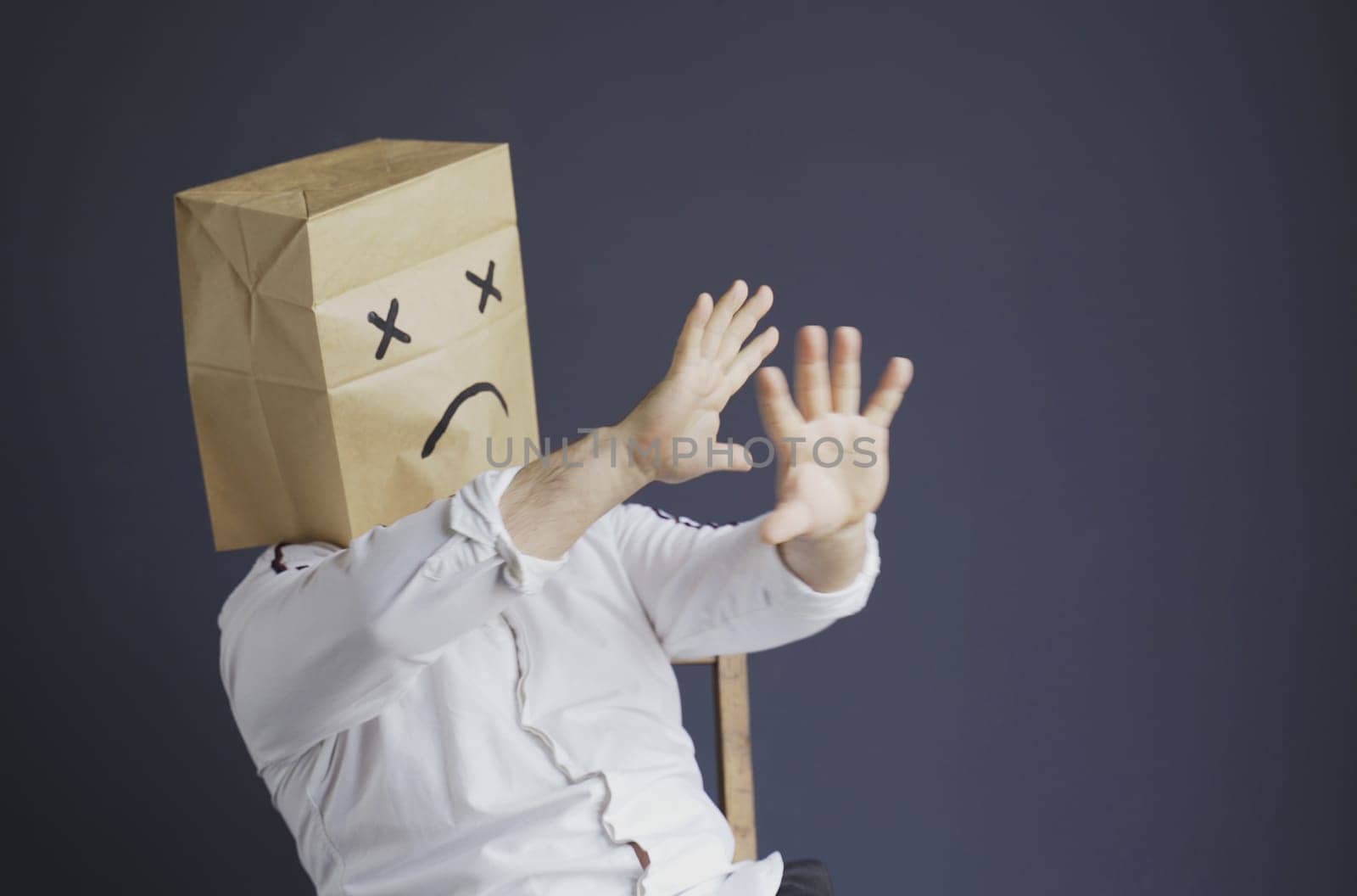 A sad man in a white shirt with a bag on his head, with a sad emoticon drawn, is afraid, defends himself by putting his hands forward. Emotions and gestures.