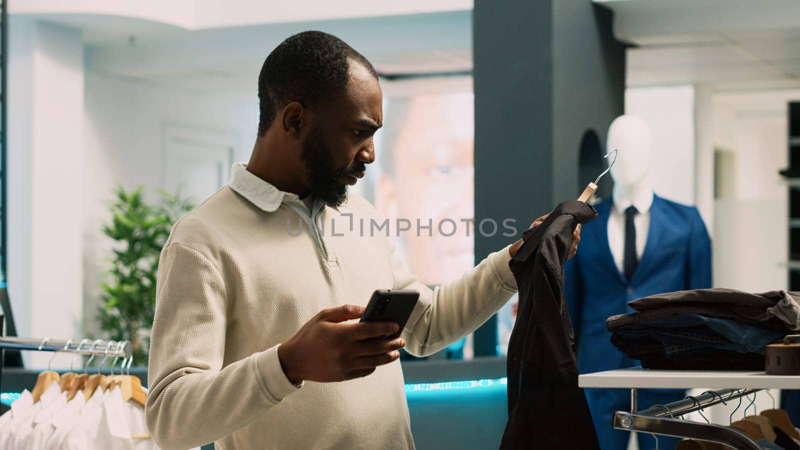 Male shopper trying to find clothes from online website, checking stock in modern clothing store. Young shopaholic man looking to buy formal or casual wear, commercial activity. Handheld shot.