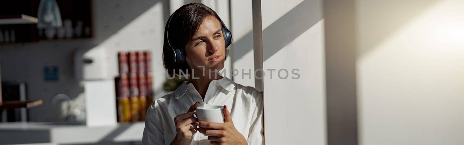 Business woman in headphones holding a cup of coffee while standing near window at office by Yaroslav_astakhov