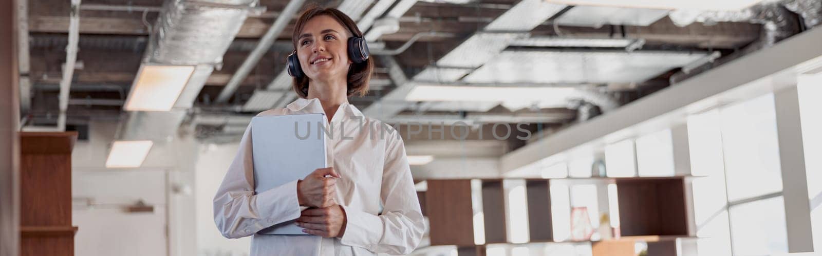 Attractive business woman in headphones with laptop standing in modern office by Yaroslav_astakhov