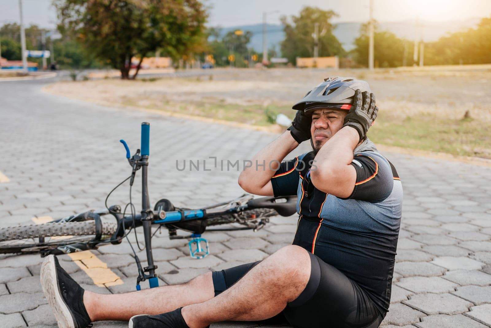 Cyclist on the pavement with a headache. Concept of cyclist suffering with migraine. Male cyclist sitting with headache by isaiphoto