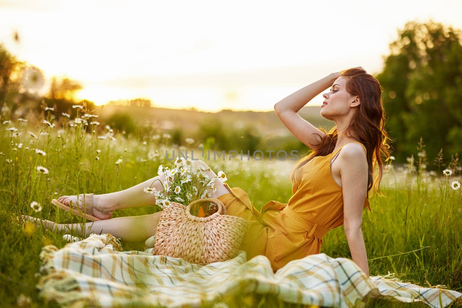 a beautiful woman in an orange dress sits on a plaid in a chamomile field and straightens her hair. High quality photo