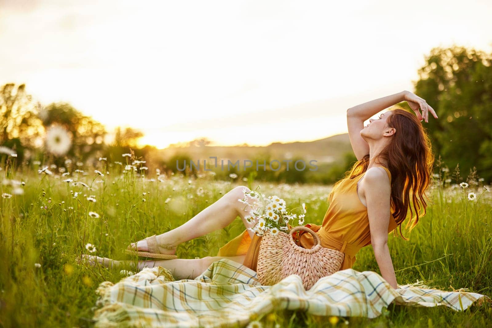 a beautiful woman in an orange dress sits on a plaid in a chamomile field and straightens her hair. High quality photo