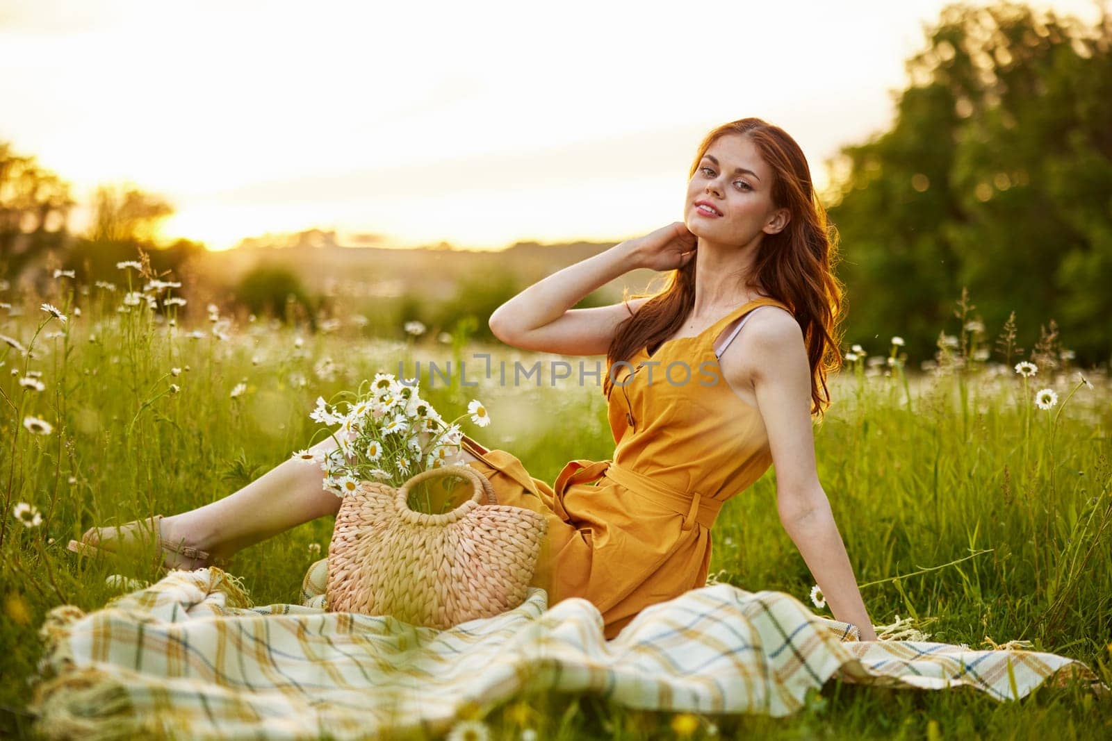 beautiful, happy woman in an orange dress is resting sitting on a plaid in a chamomile field. High quality photo