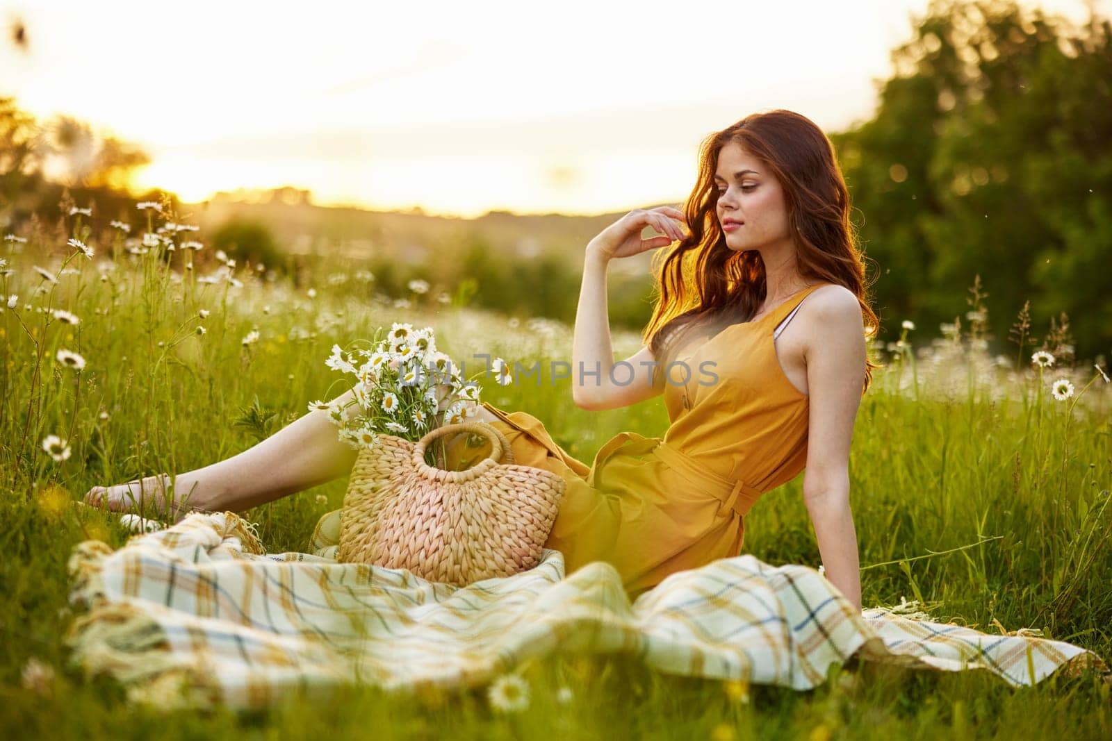 a beautiful woman in an orange dress sits on a plaid in a chamomile field and straightens her hair. High quality photo