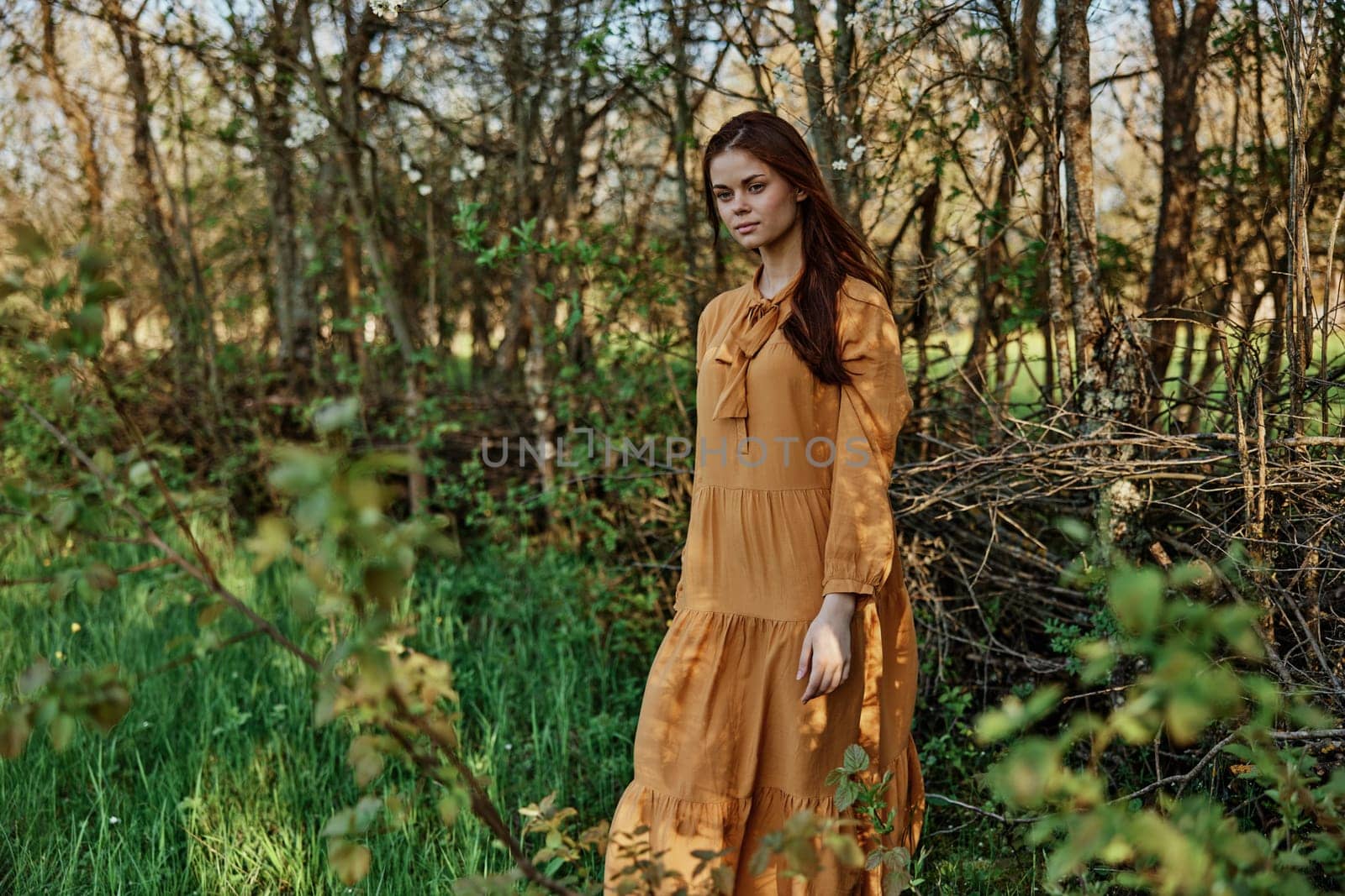 a beautiful woman walks in the shade near the trees, dressed in a long orange dress, enjoying the weather and the weekend, posing relaxed. The theme of privacy with nature. High quality photo