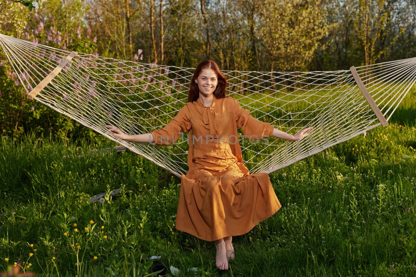 horizontal photo of a beautiful, red-haired woman lying in a hammock enjoying a rest in a long orange dress, on a warm summer day, smiling happily looking at the camera by Vichizh