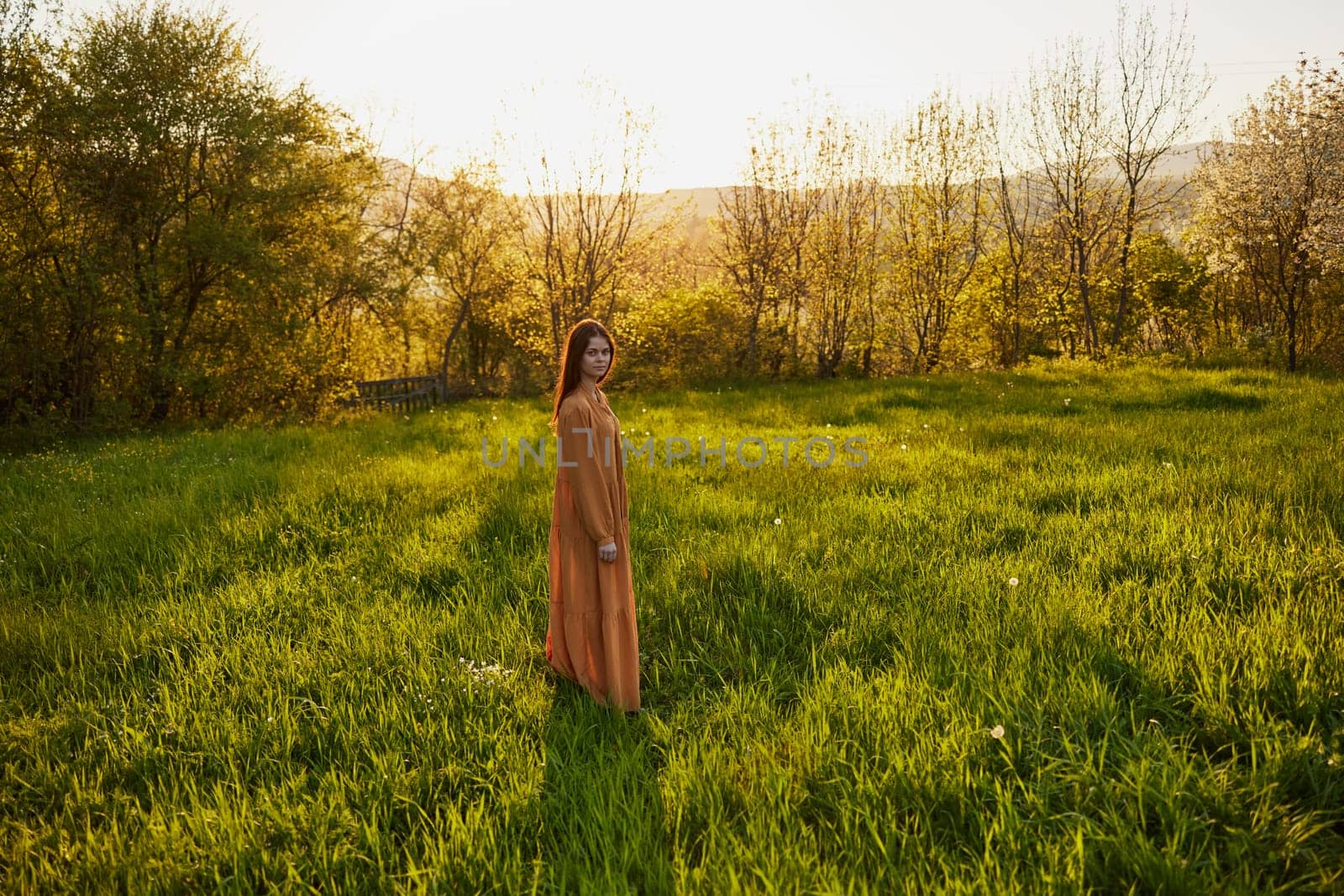 an attractive, slender, red-haired woman stands in a wide green field during sunset in a long orange dress enjoying unity with nature and relaxation by Vichizh