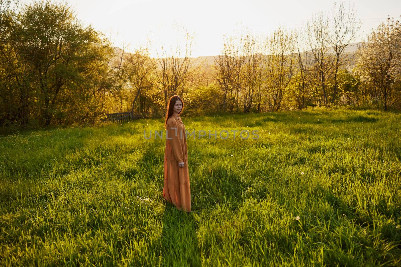 an attractive, slender, red-haired woman stands in a wide green field during sunset in a long orange dress enjoying unity with nature and relaxation. High quality photo