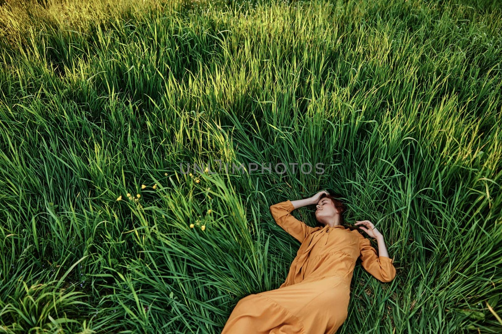 a sweet, calm woman in an orange dress lies in a green field with her hands under her head enjoying silence and peace. Horizontal photo taken from above. High quality photo