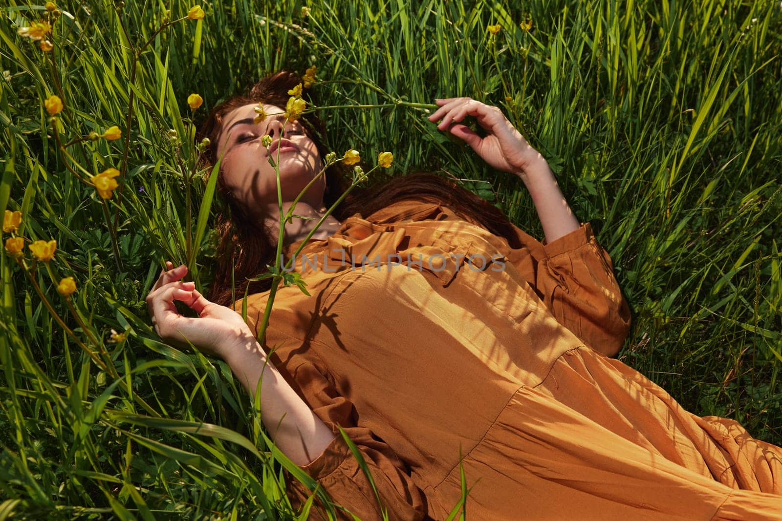 a calm woman with long red hair lies in a green field with yellow flowers, in an orange dress with her eyes closed, spreading her arms to the sides, enjoying peace and recuperating. High quality photo