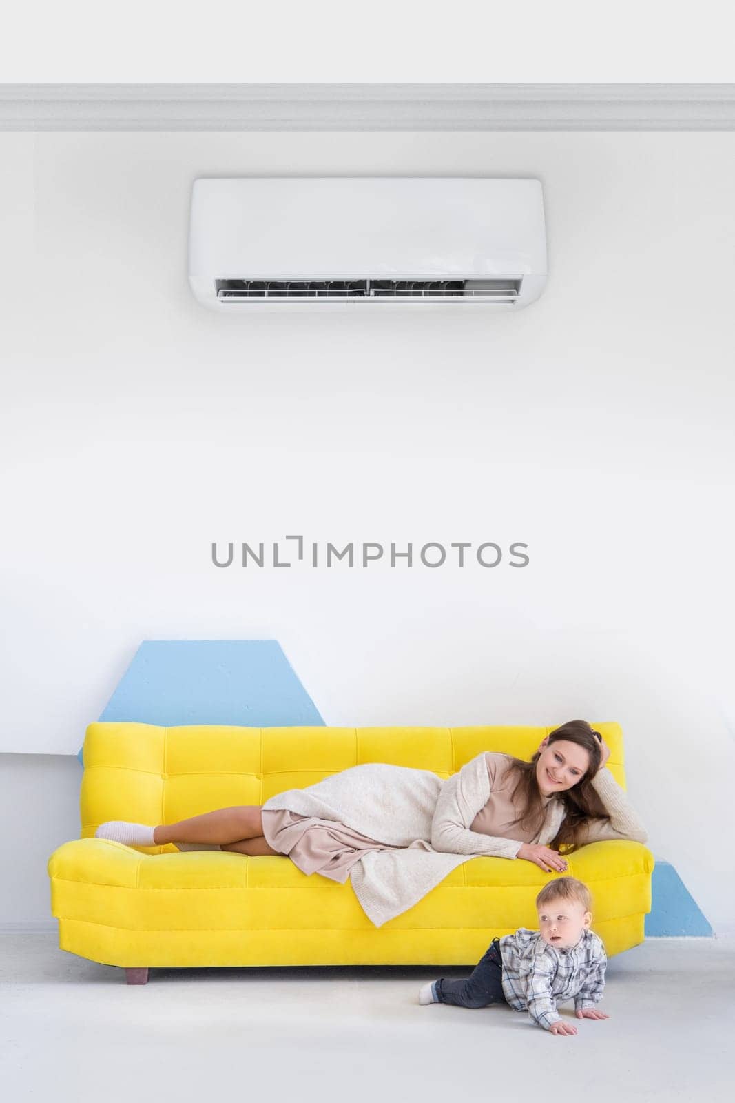 Young caucasian woman with child lies on a yellow sofa in bright interior with split air conditioner.