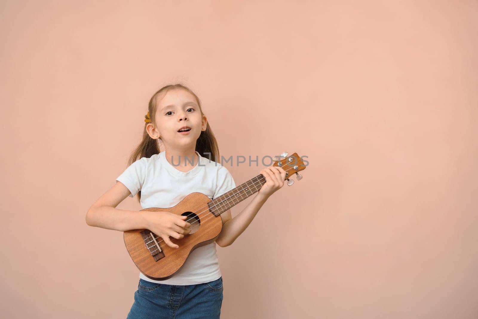 Kid girl in a white T-shirt with a ukulele on a pink background, copy space.