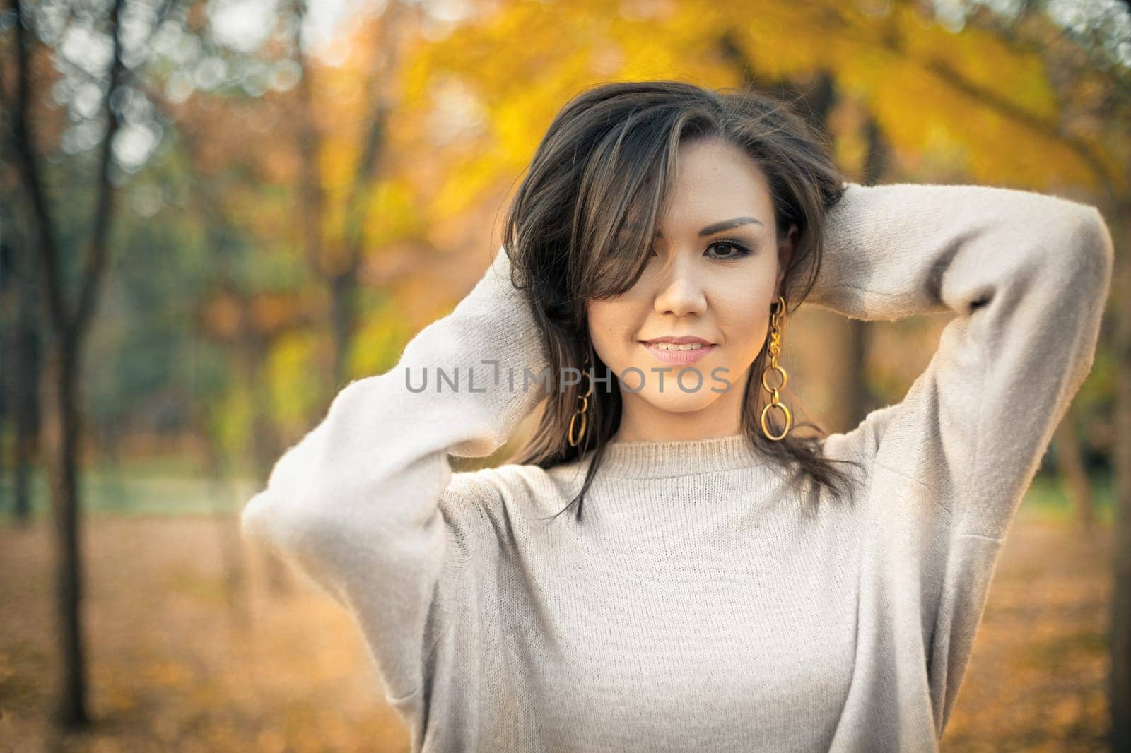 Young beautiful mixed race woman 30s in gray sweater posing in autumn park looking at camera.