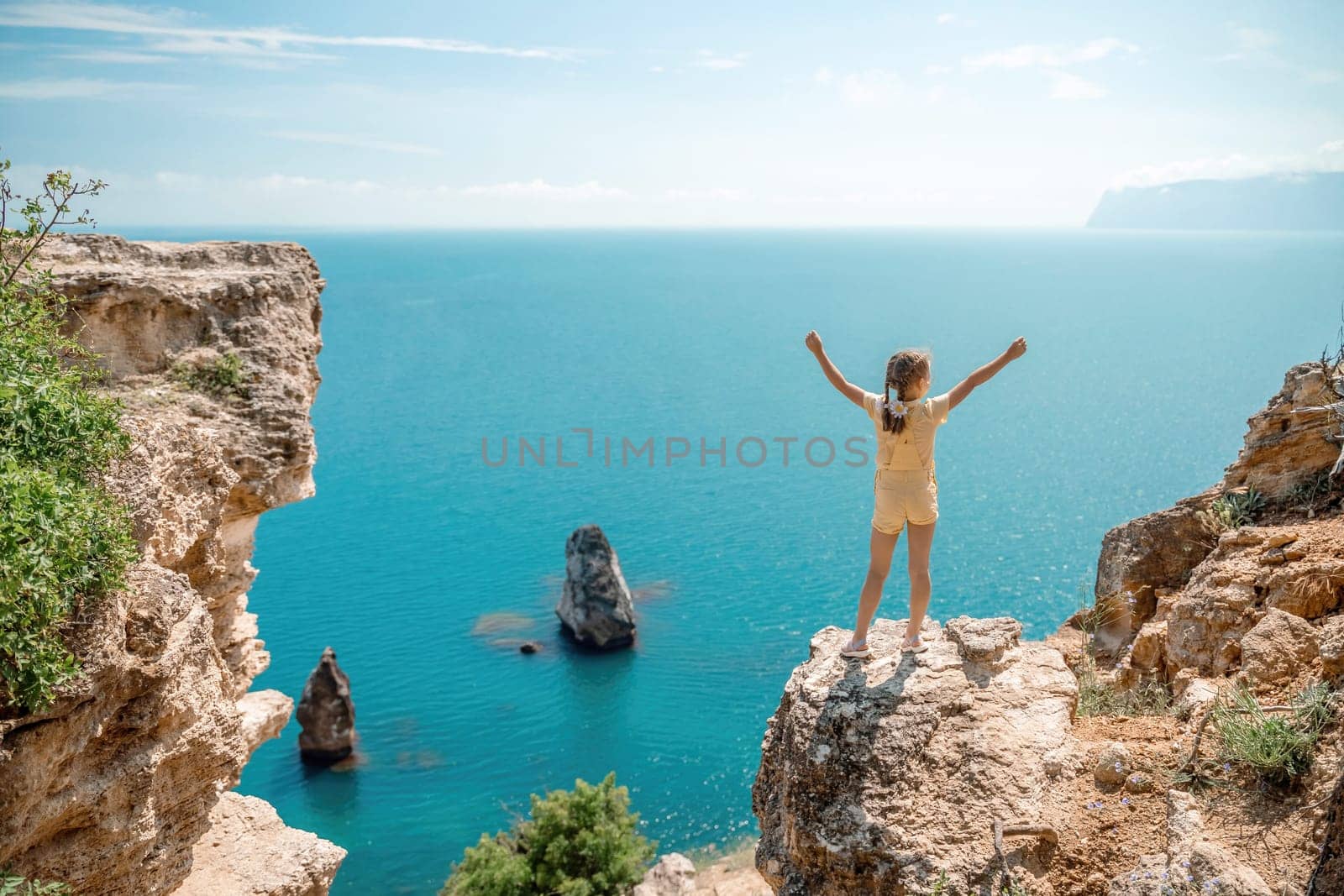 Happy girl stands on a rock high above the sea, wearing a yellow jumpsuit and sporting braided hair, depicting the idea of a summer vacation by the sea. by Matiunina