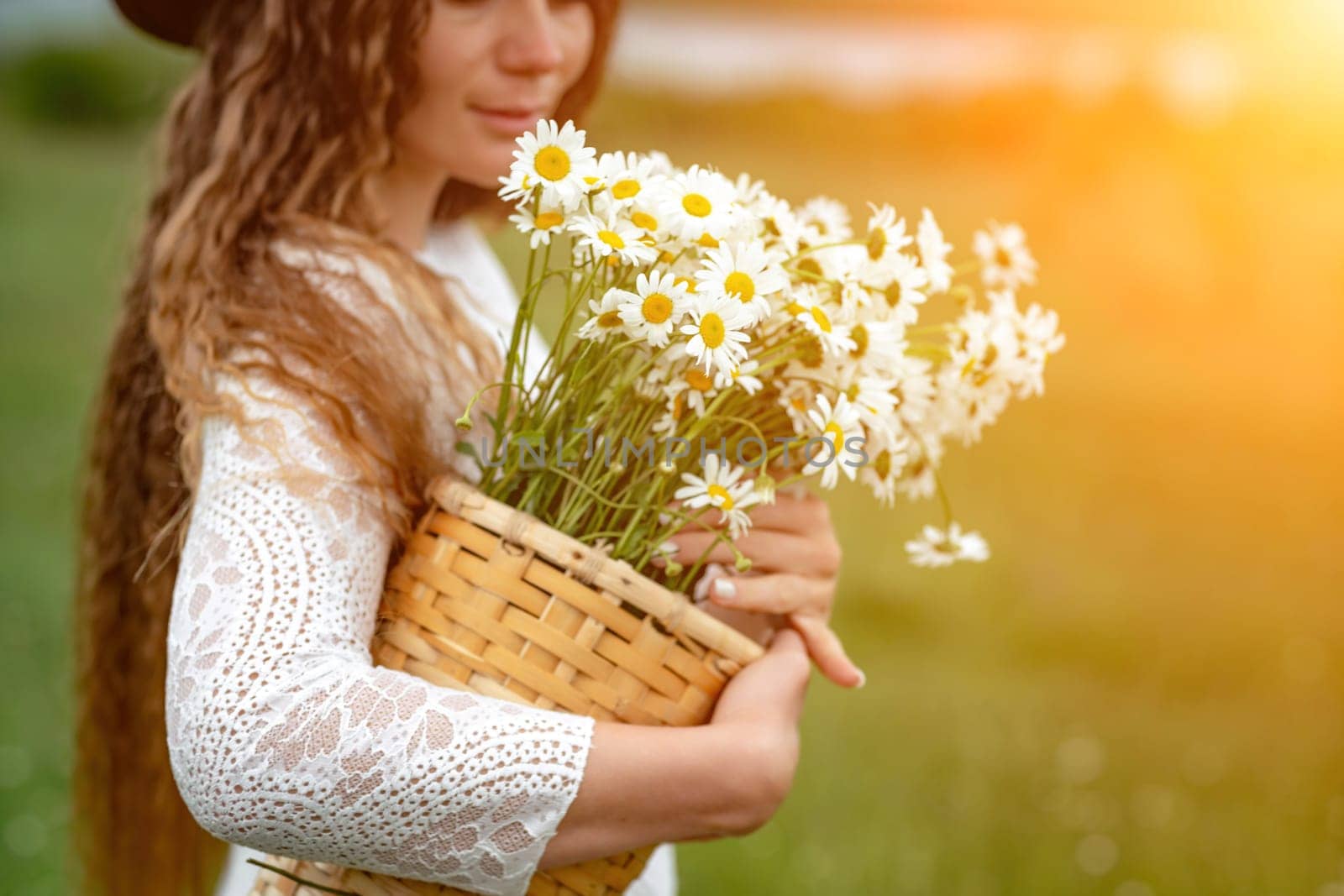 A middle-aged woman in a white dress and brown hat holds a lA middle-aged woman in a white dress and brown hat holds a basket in her hands with a large bouquet of daisies.arge bouquet of daisies in her hands. Wildflowers for congratulations.