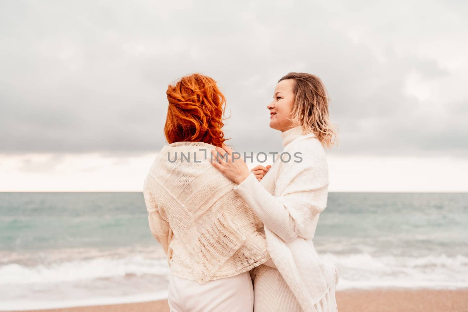 Women sea walk friendship spring. Two girlfriends, redhead and blonde, middle-aged walk along the sandy beach of the sea, dressed in white clothes. Against the backdrop of a cloudy sky and the winter sea. Weekend concept. by Matiunina