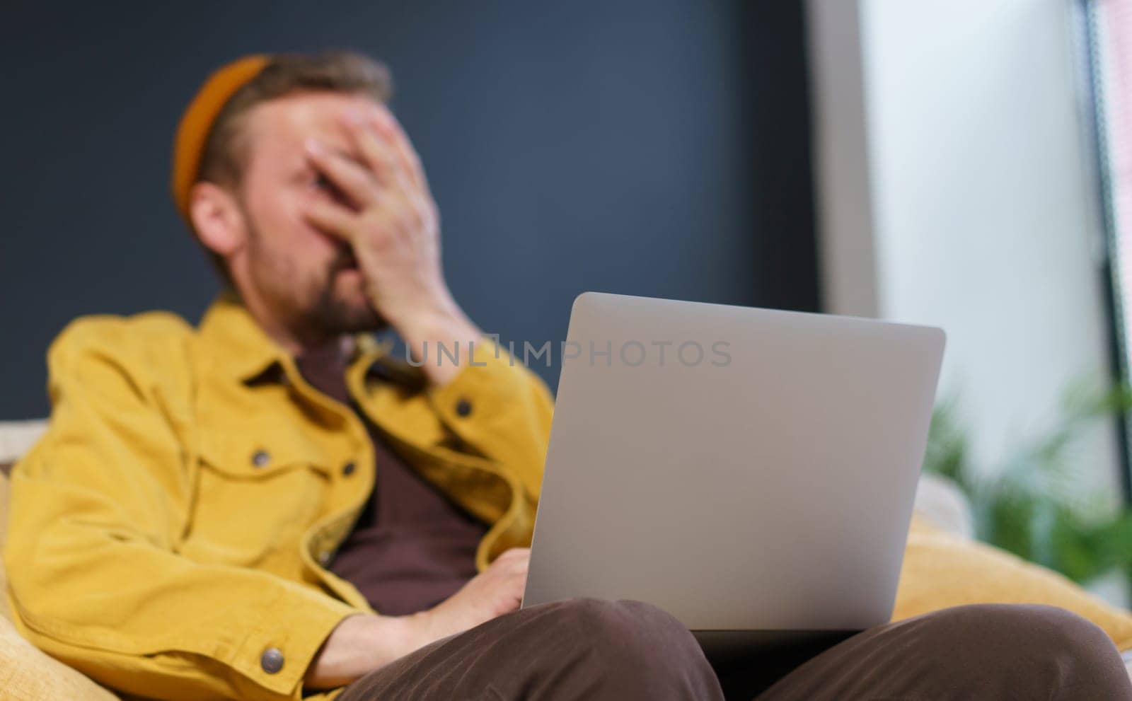 Man sitting on sofa with laptop, showing facepalm gesture with hand on face. Focus is on laptop, which suggests that the man may be experiencing frustration or disappointment related to the technology. High quality photo