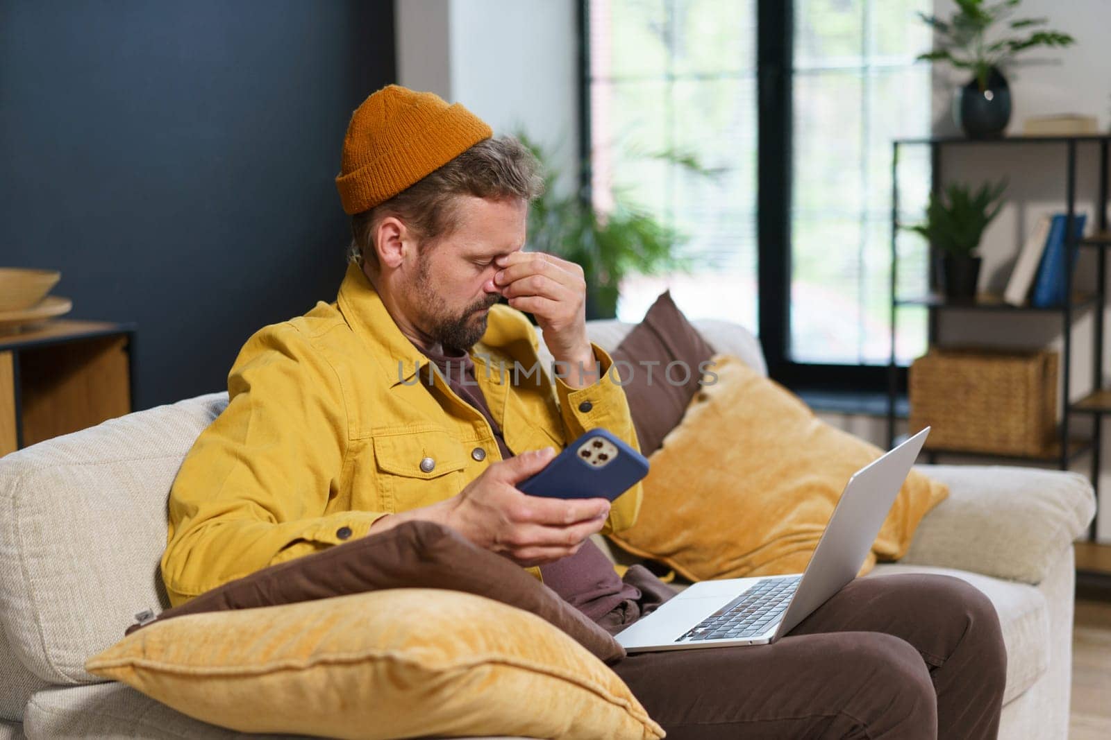 Tired man in feeling headache while holding hand on head with closed eyes. He sit home with mobile phone in hand and laptop on knees, suggesting he overwhelmed with work from home and multitasking. . High quality photo