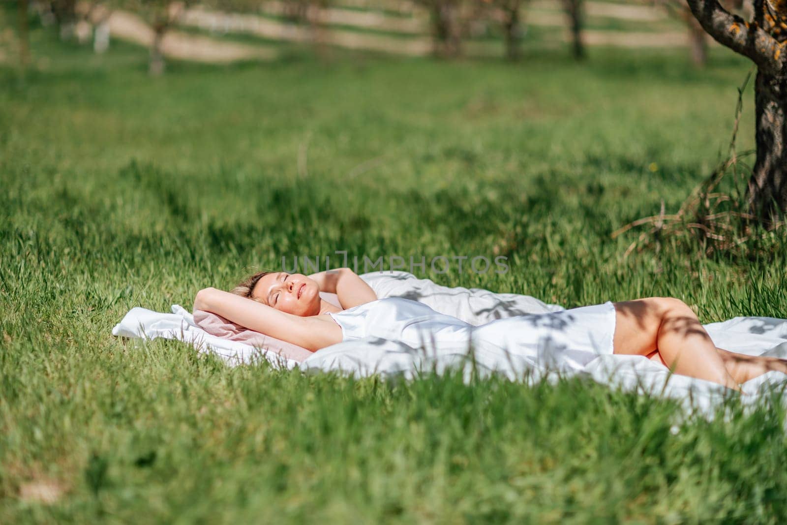 woman sleeps on a white bed in the fresh spring grass in the garden. Dressed in a blue nightgown. by Matiunina