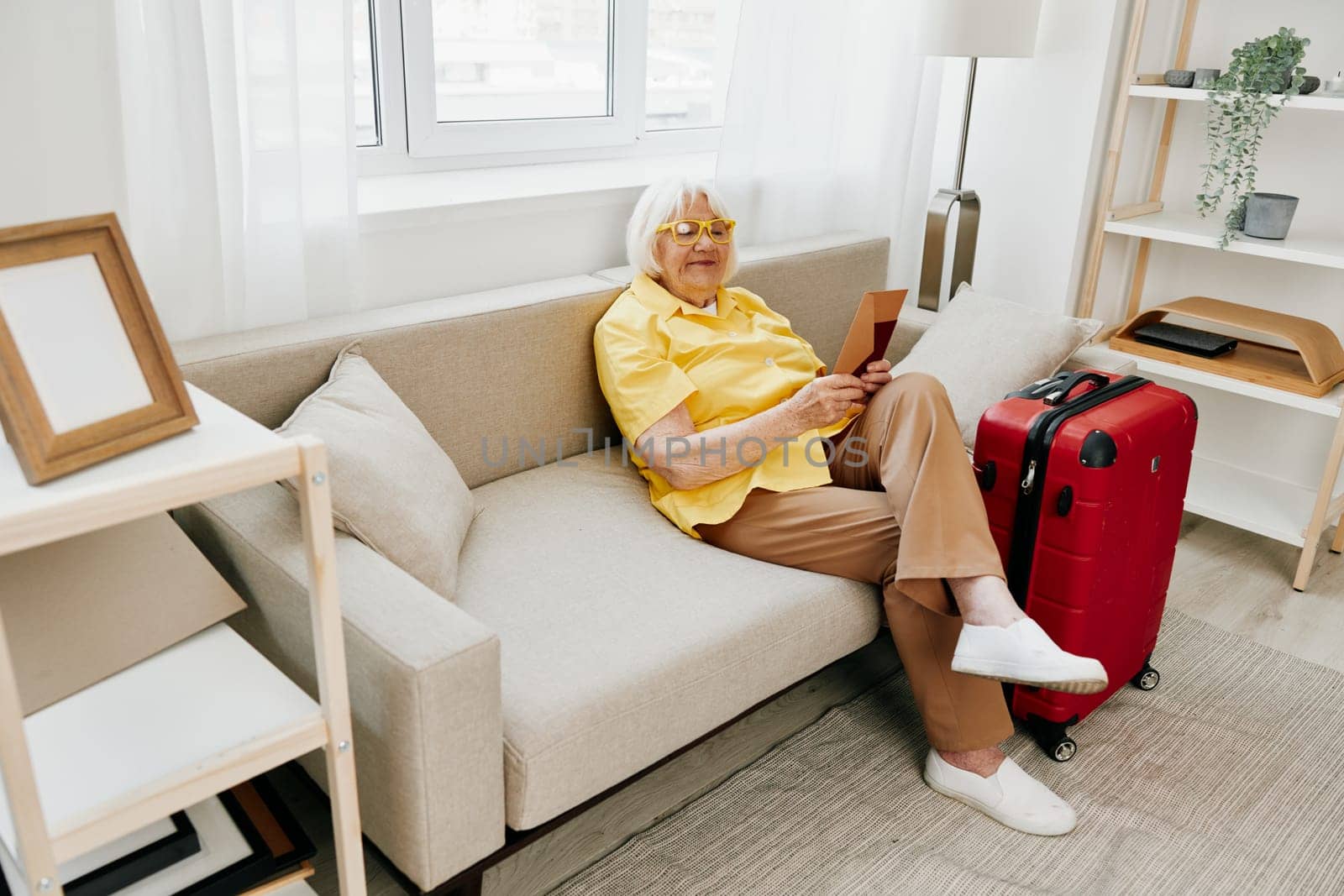 Happy senior woman with passport and travel ticket packed a red suitcase, vacation and health care. Smiling old woman joyfully sitting on the sofa before the trip raised her hands up in joy. High quality photo