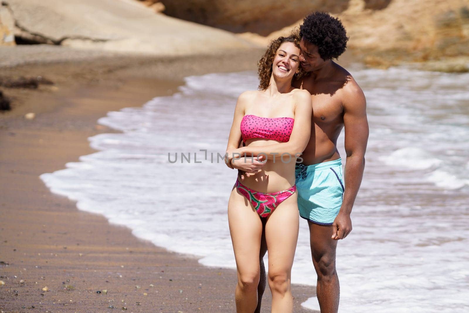 Cheerful young multiracial man and woman smiling happily and embracing each other while standing together on sandy shore during vacation