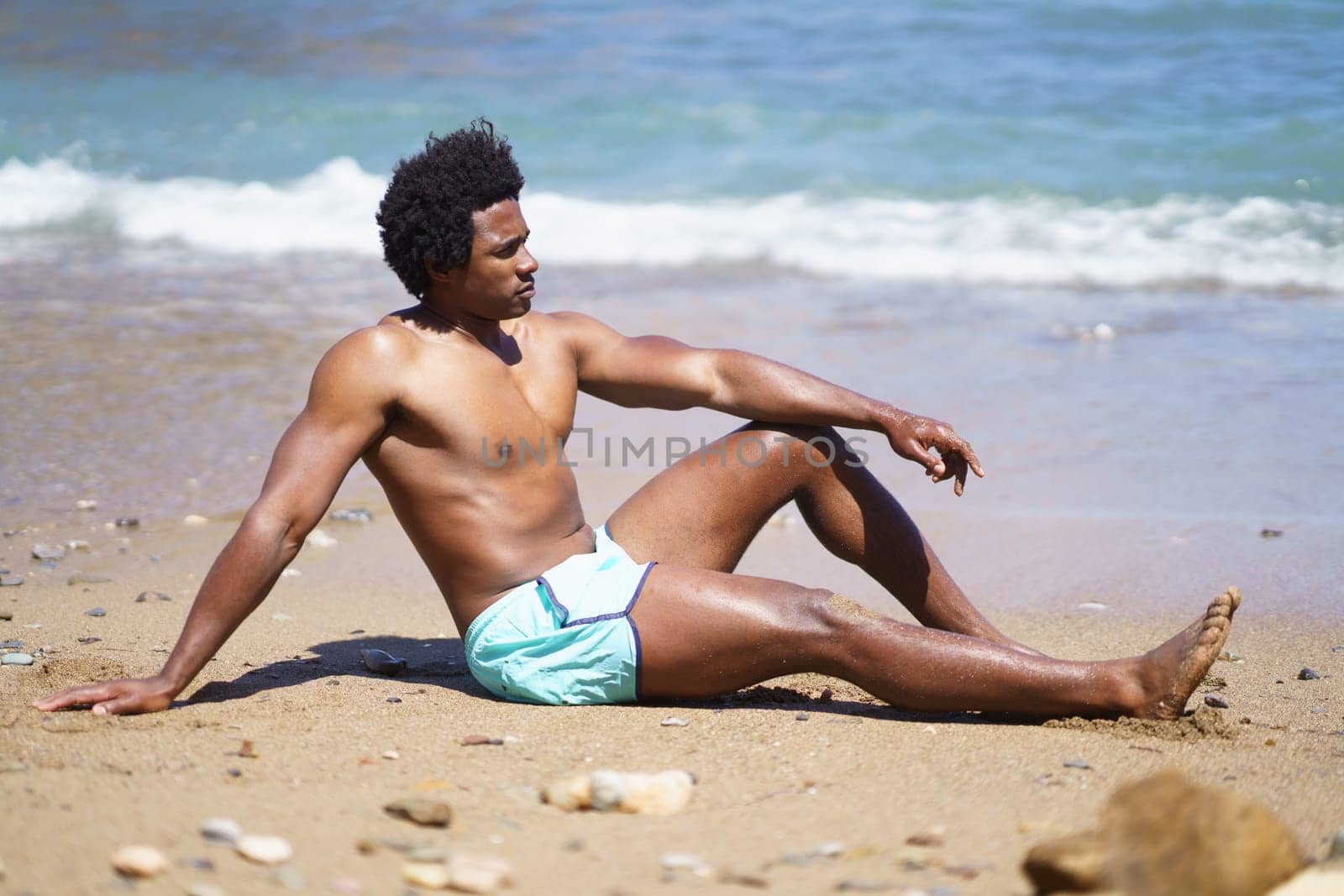 Full body side view of African American male relaxing on sandy beach with arm on knee while looking away, and leaning on hand near waving sea