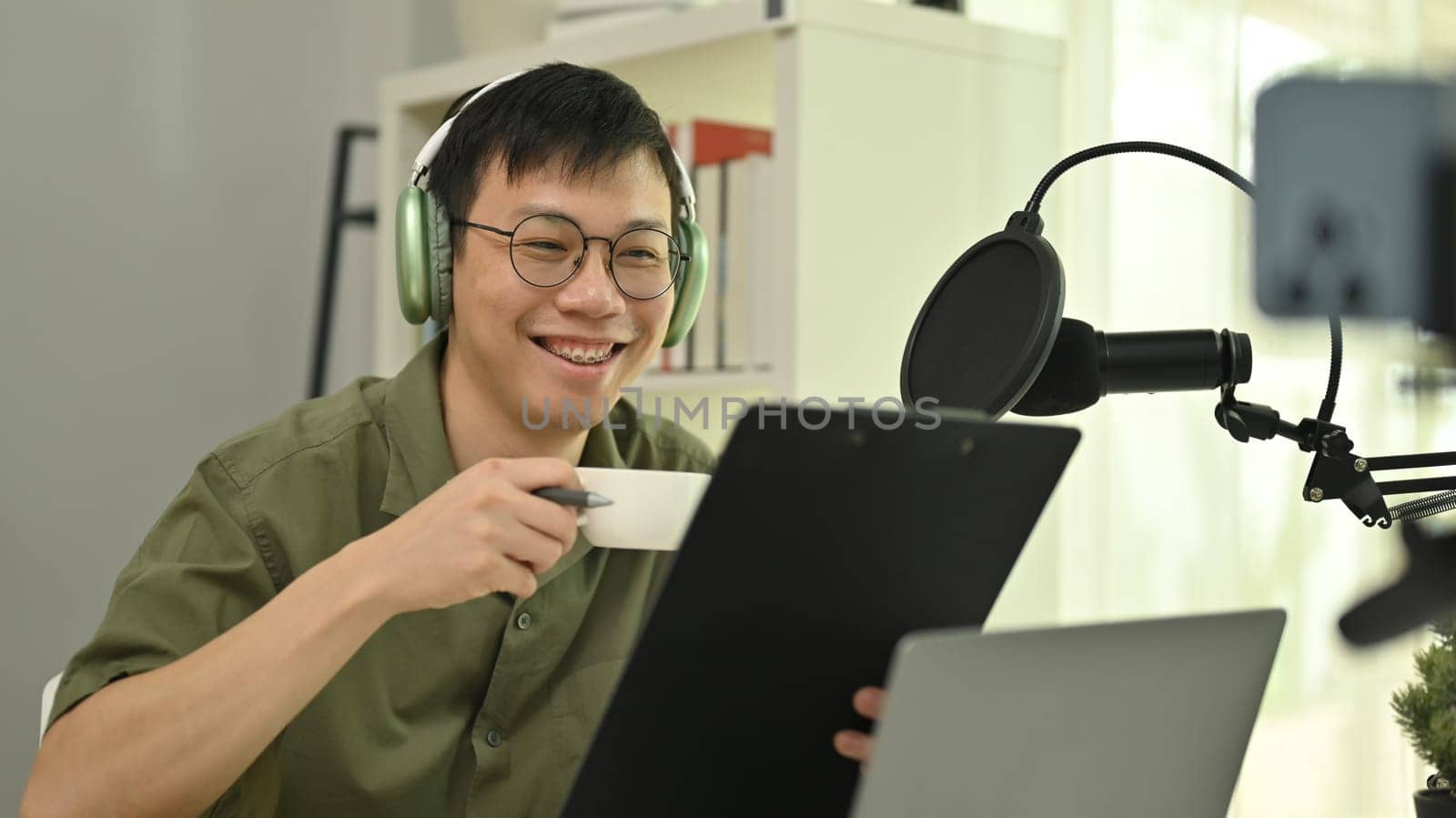 Young male podcaster using a microphone and laptop computer recording audio for channel in home studio by prathanchorruangsak