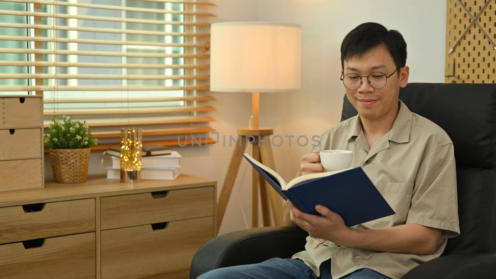 Calm asian man wearing glasses sitting on sofa and reading book. Leisure and people concept.