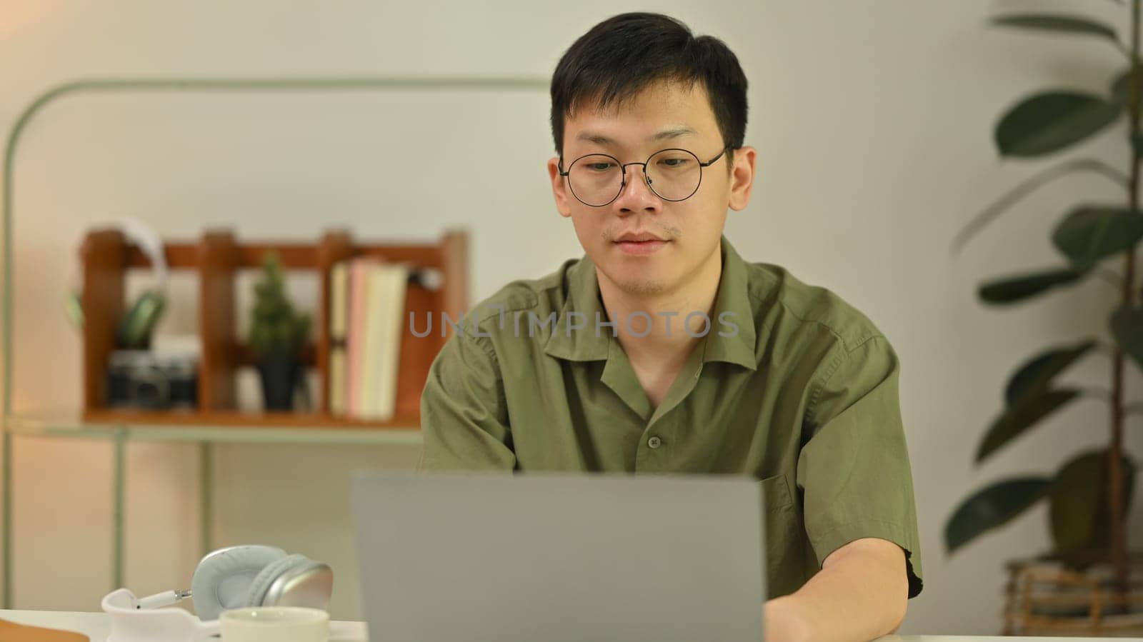 Millennial asian man in glasses looking at laptop screen and typing on keyboard. Technology, freelance and remote work concept.