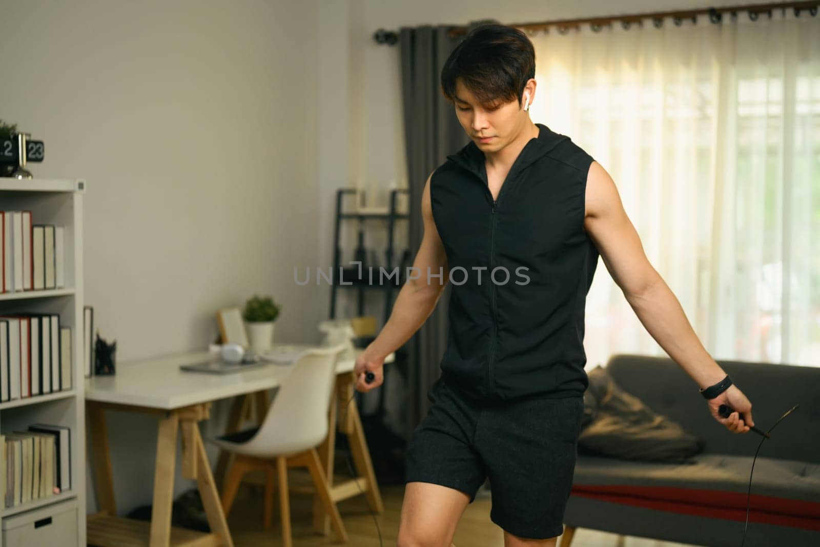 Handsome male athlete in sportswear exercising with jumping rope in modern living room by prathanchorruangsak