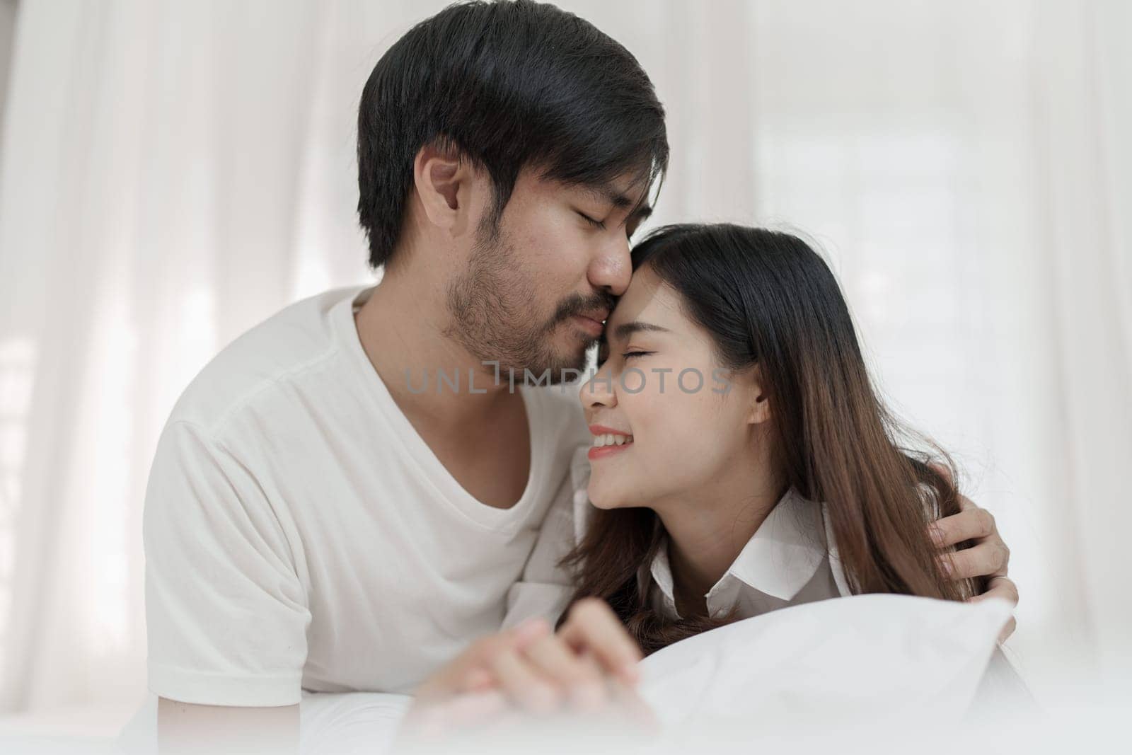 Asian Romantic couple in bed enjoying sensual foreplay Happy sensual young couple lying in bed together. Beautiful loving couple kissing in bed. by Manastrong