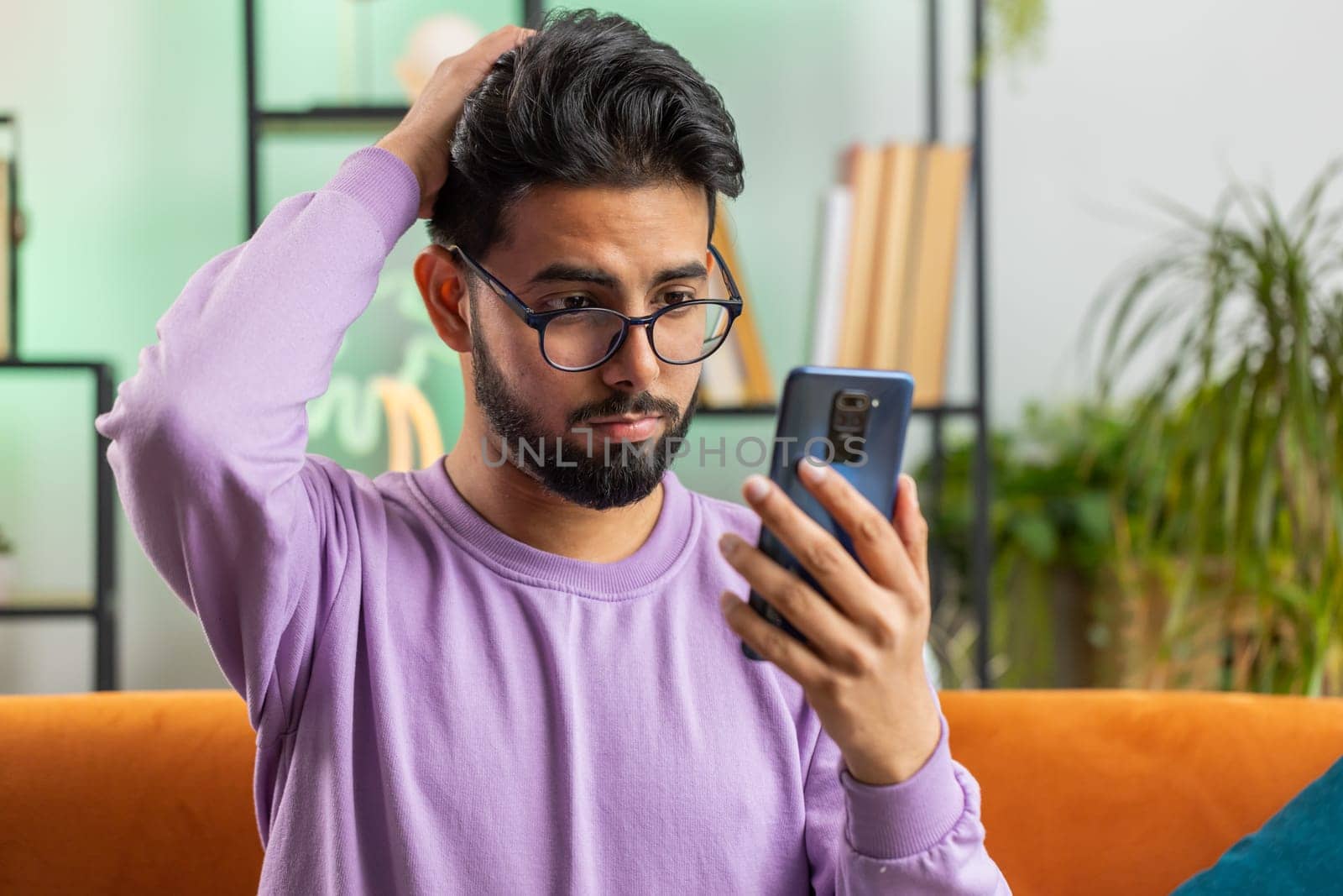Sad displeased indian man use smartphone typing browsing, loses becoming surprised sudden lottery results, bad news, fortune loss, fail, deadline, virus. Young hindu guy at home living room on couch