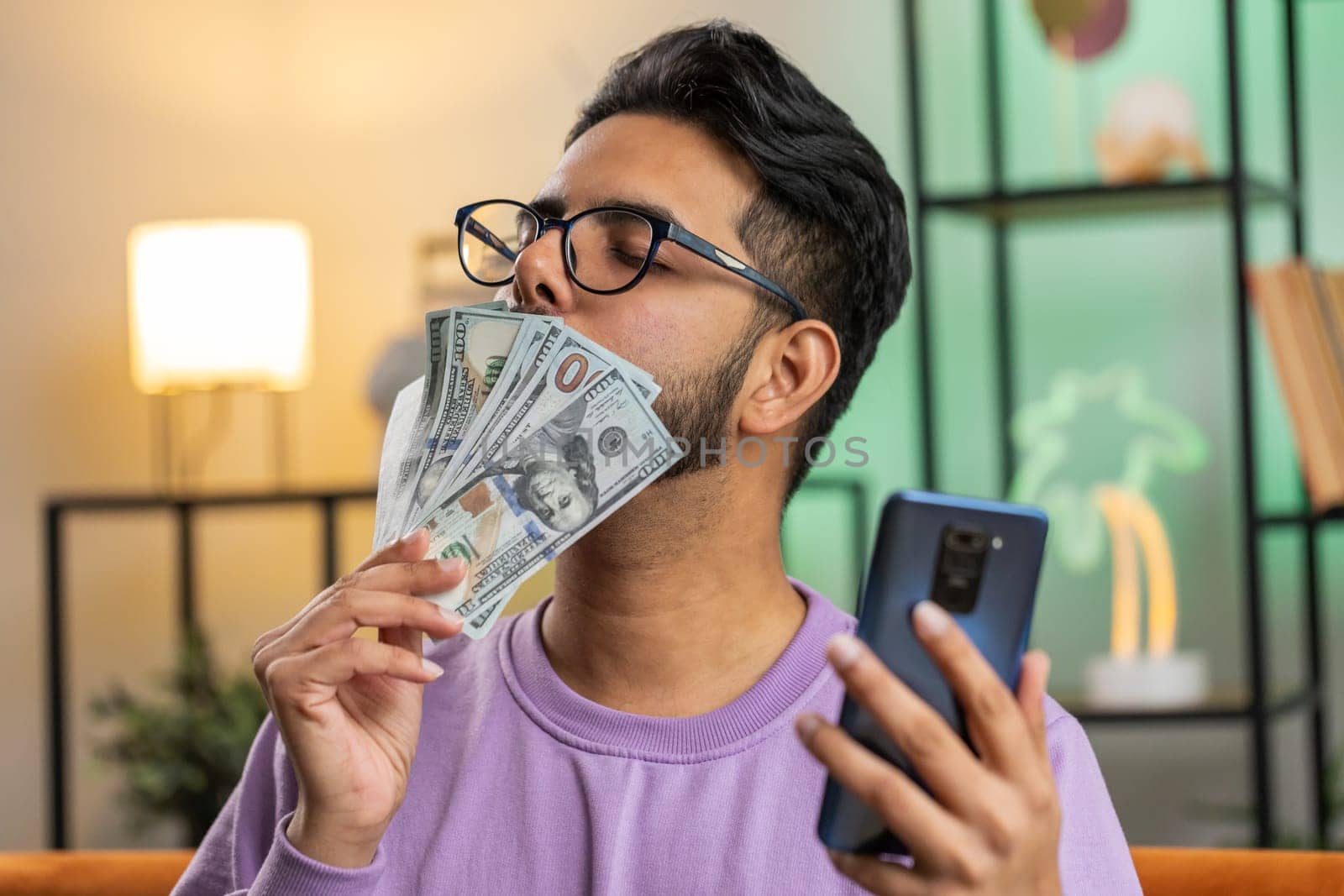 Planning family budget. Smiling indian man counting money cash, use smartphone calculate domestic bills at home room. Joyful hindu guy satisfied of income and saves money for planned vacation, gifts