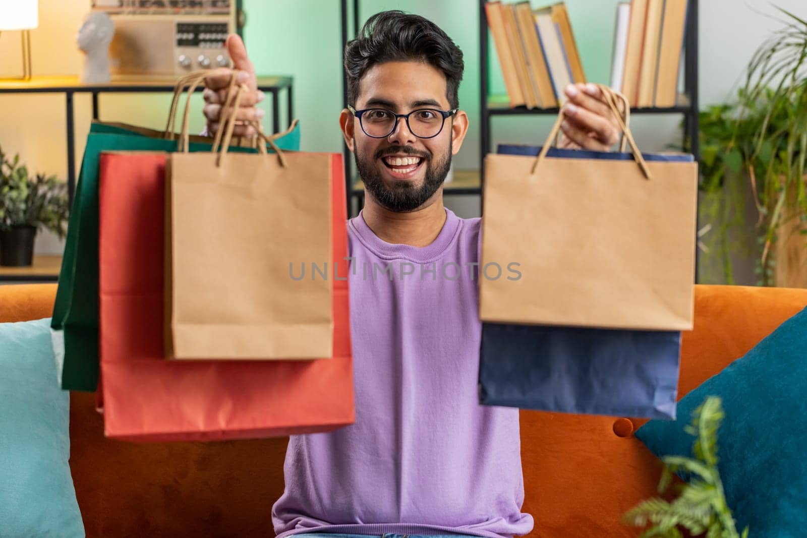 Young indian man happy shopaholic consumer came back home after shopping sale with bags. Portrait of hindu guy satisfied received parcels purchase from online order at home apartment room on sofa