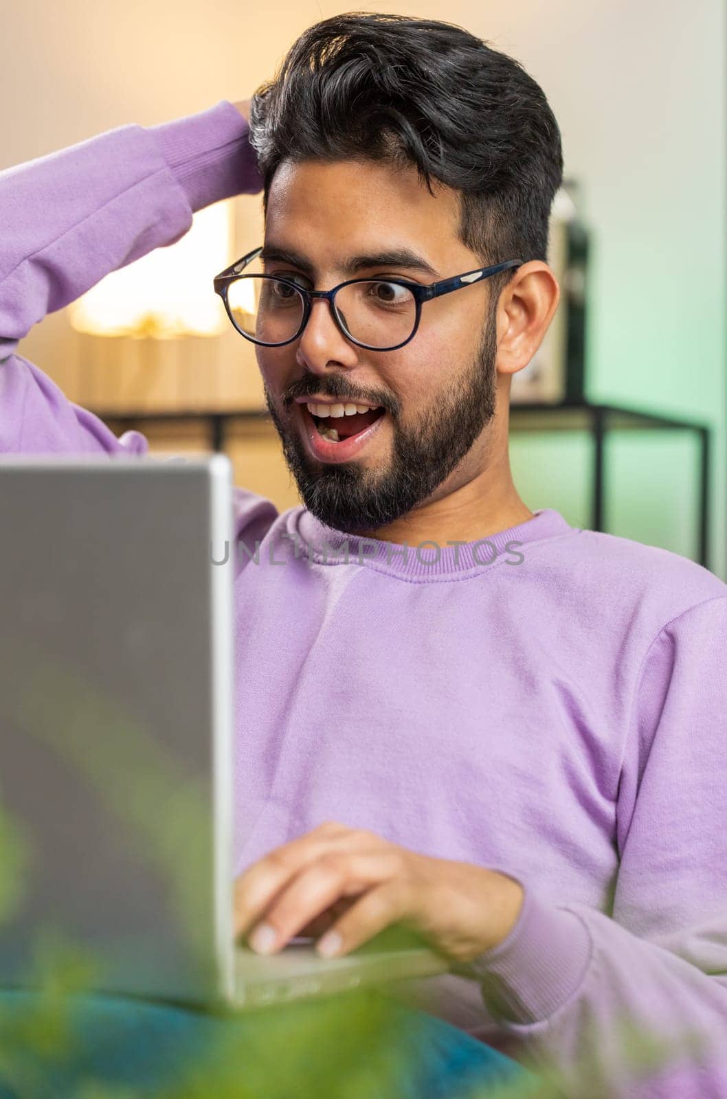 Surprised indian man guy using laptop computer, receive mail good news message, shocked by sudden victory celebrate lottery jackpot win purchases online shopping at home apartment on sofa. Lifestyles