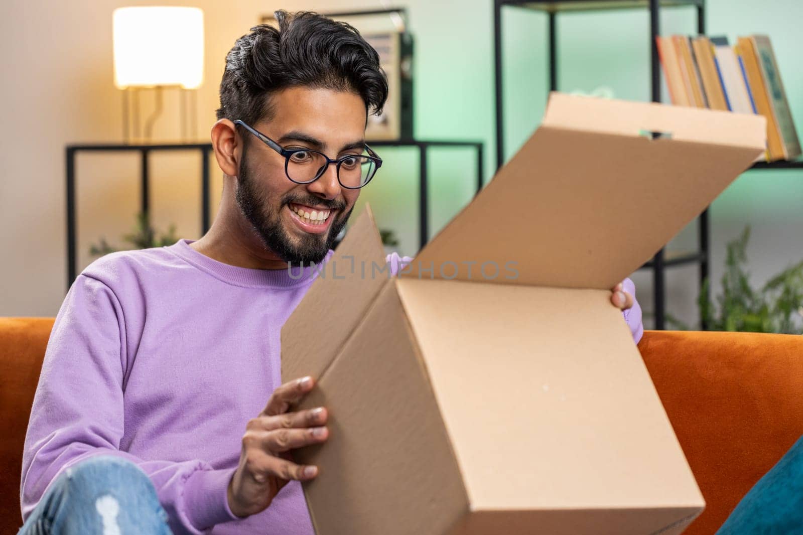 Happy indian man unpacking delivery parcel sitting at home apartment. Smiling satisfied hindu guy shopper, online shop customer opening cardboard box receiving purchase gift by fast postal shipping
