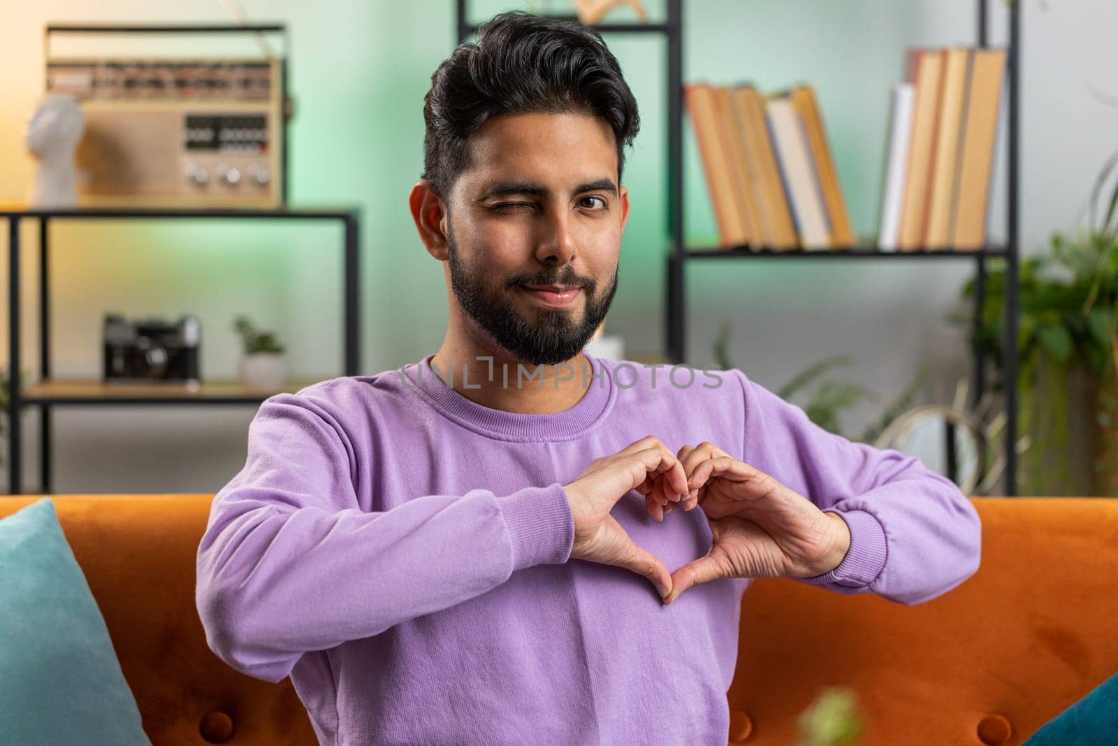 I love you. Happy indian man at home living room couch makes symbol of love, showing heart sign to camera, express romantic feelings express sincere positive feelings. Charity, gratitude, donation