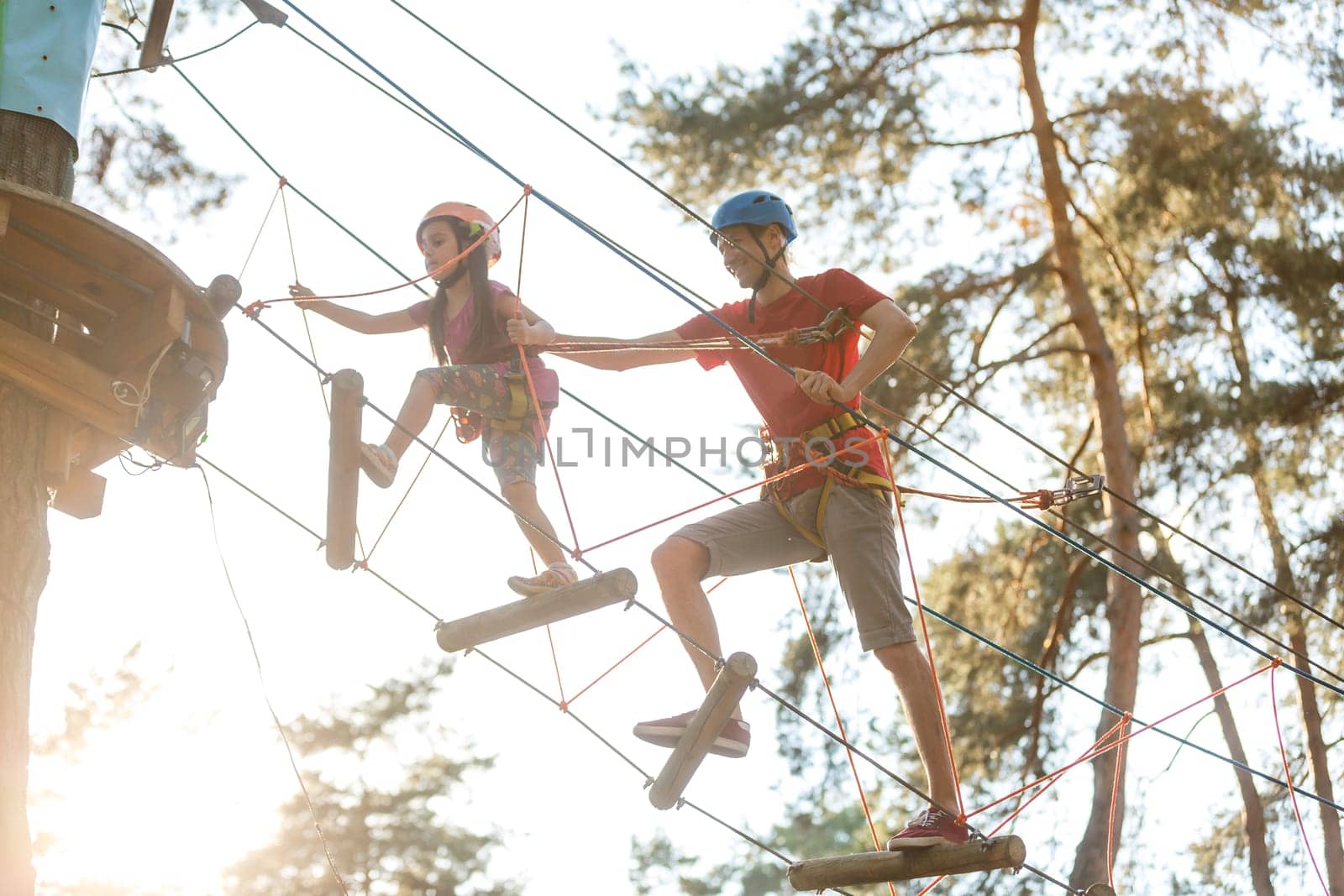 Young father helps his daughter on the obstacle in the rope park.