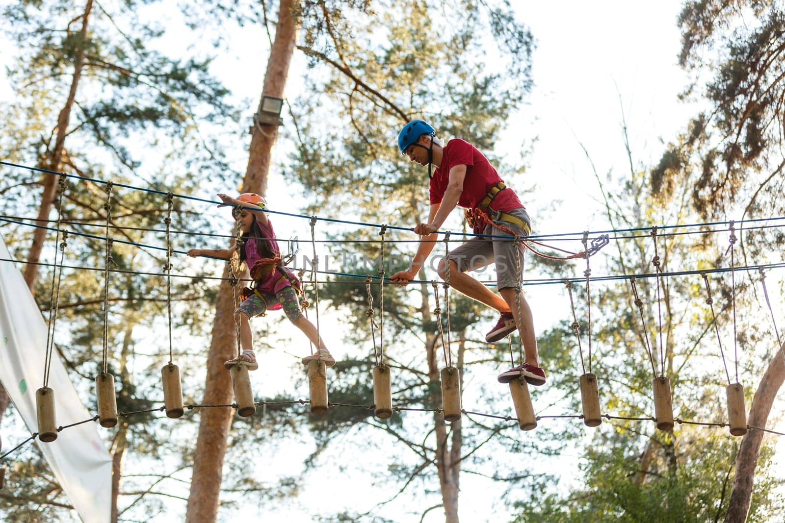 Young father helps his daughter on the obstacle in the rope park.