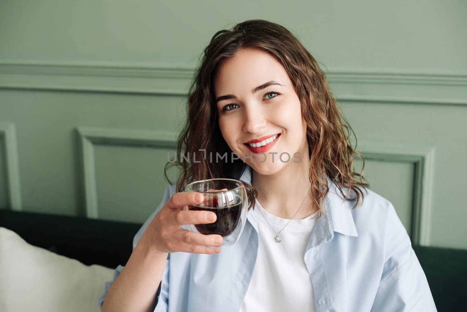 Happy young woman housewife enjoying a cozy break with hot tea on the sofa at home. Smiling lady relaxing with a warm drink and looking at the camera in a close-up indoor portrait
