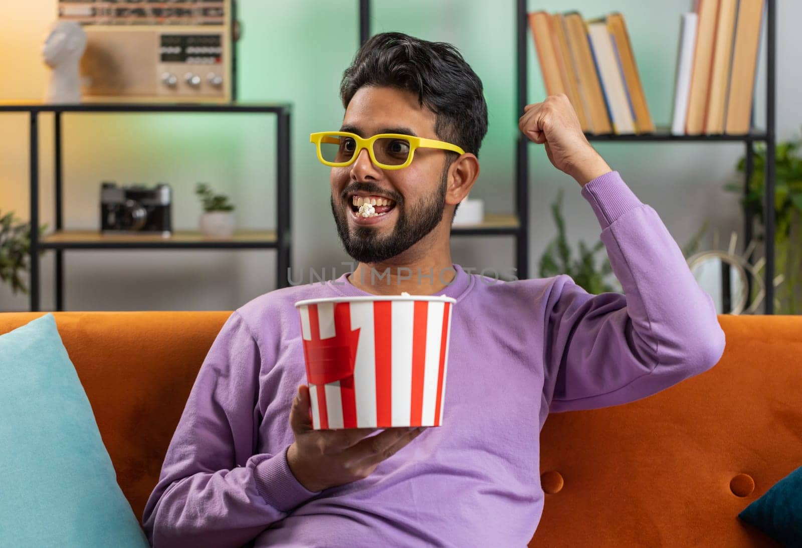 Excited indian man in 3D glasses sits on sofa eating popcorn snacks and watching interesting TV serial sport game, film, online social media movie content at home apartment. Guy enjoying entertainment