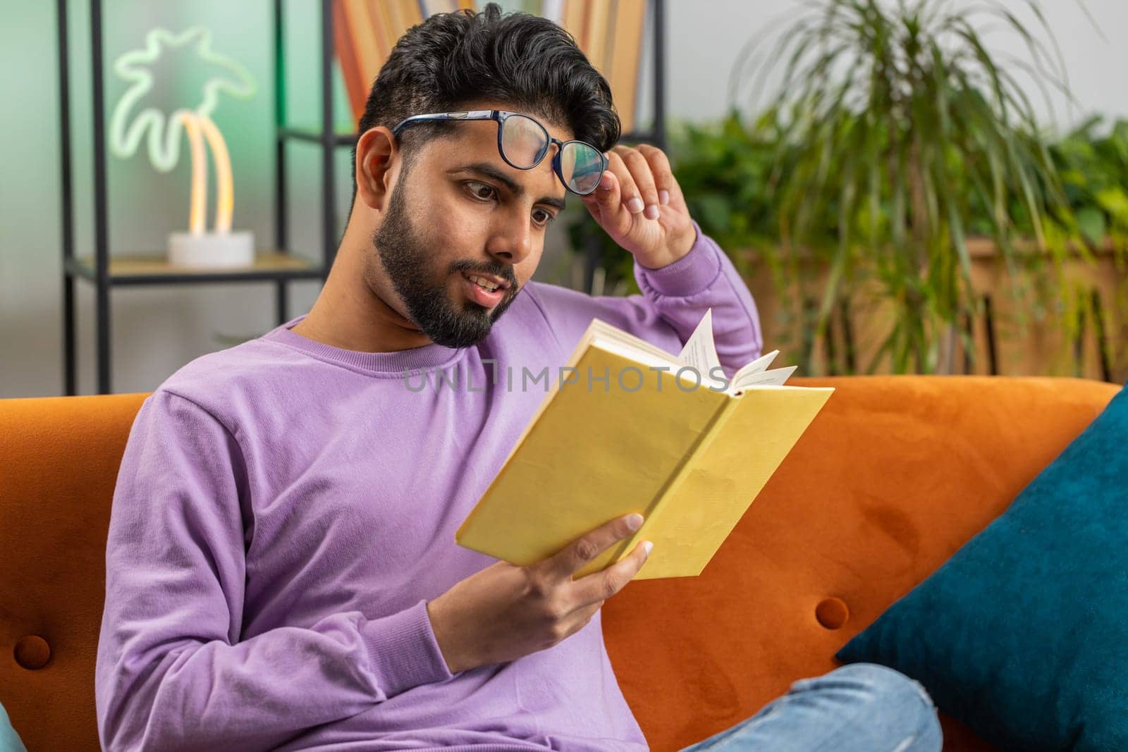 Young indian man reading interesting book, turning pages smiling enjoying literature, taking a rest on comfortable couch. Portrait of peaceful cheerful hindu guy at home apartment living room on sofa